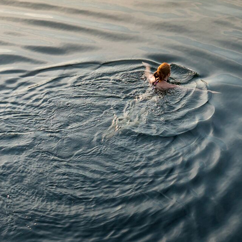 A person with light hair swims in calm water, creating wide ripples across the surface.